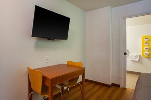 Una habitación con una televisión de pantalla plana montada en la pared, una mesa de madera con dos sillas y una puerta que da a un baño con toallero.