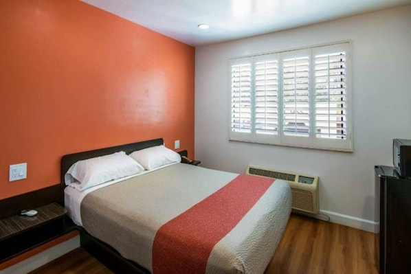 Une chambre d’hôtel avec un lit, un radiateur, une fenêtre avec des stores et un mur avec un détail décoratif rouge.