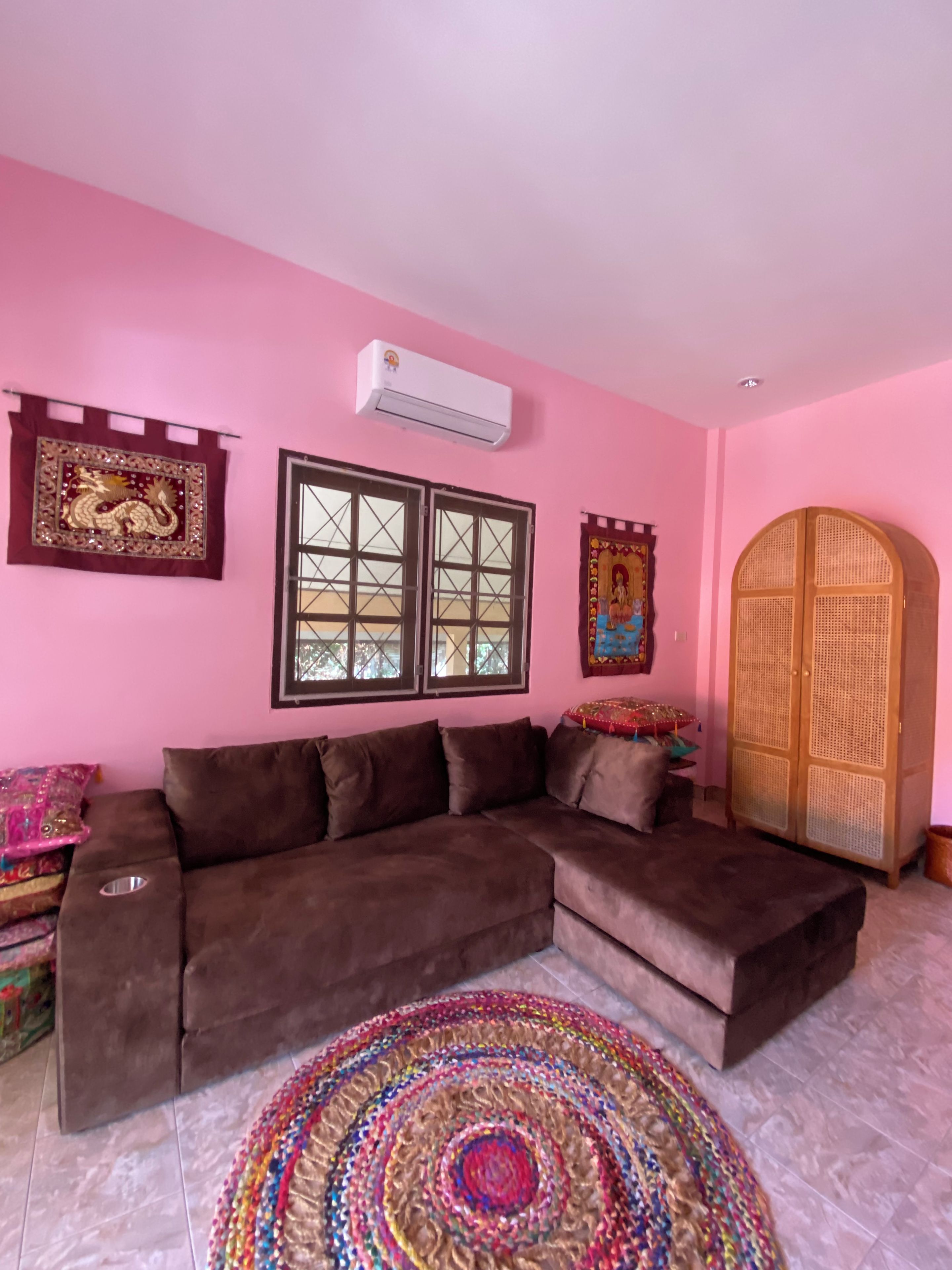 A living room with a brown sectional sofa, a colorful woven rug, a large wooden wardrobe, and decorative wall hangings.