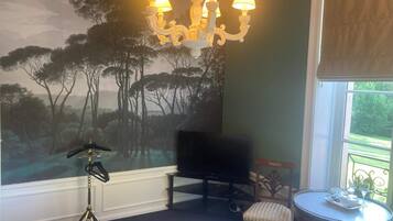 A hotel room with a bed, a chandelier, a television, a small table with a tray, and a mural of trees.