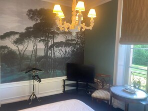 Una camera d'albergo con un letto, un lampadario, una televisione, un tavolino con un vassoio e un affresco raffigurante alberi.
