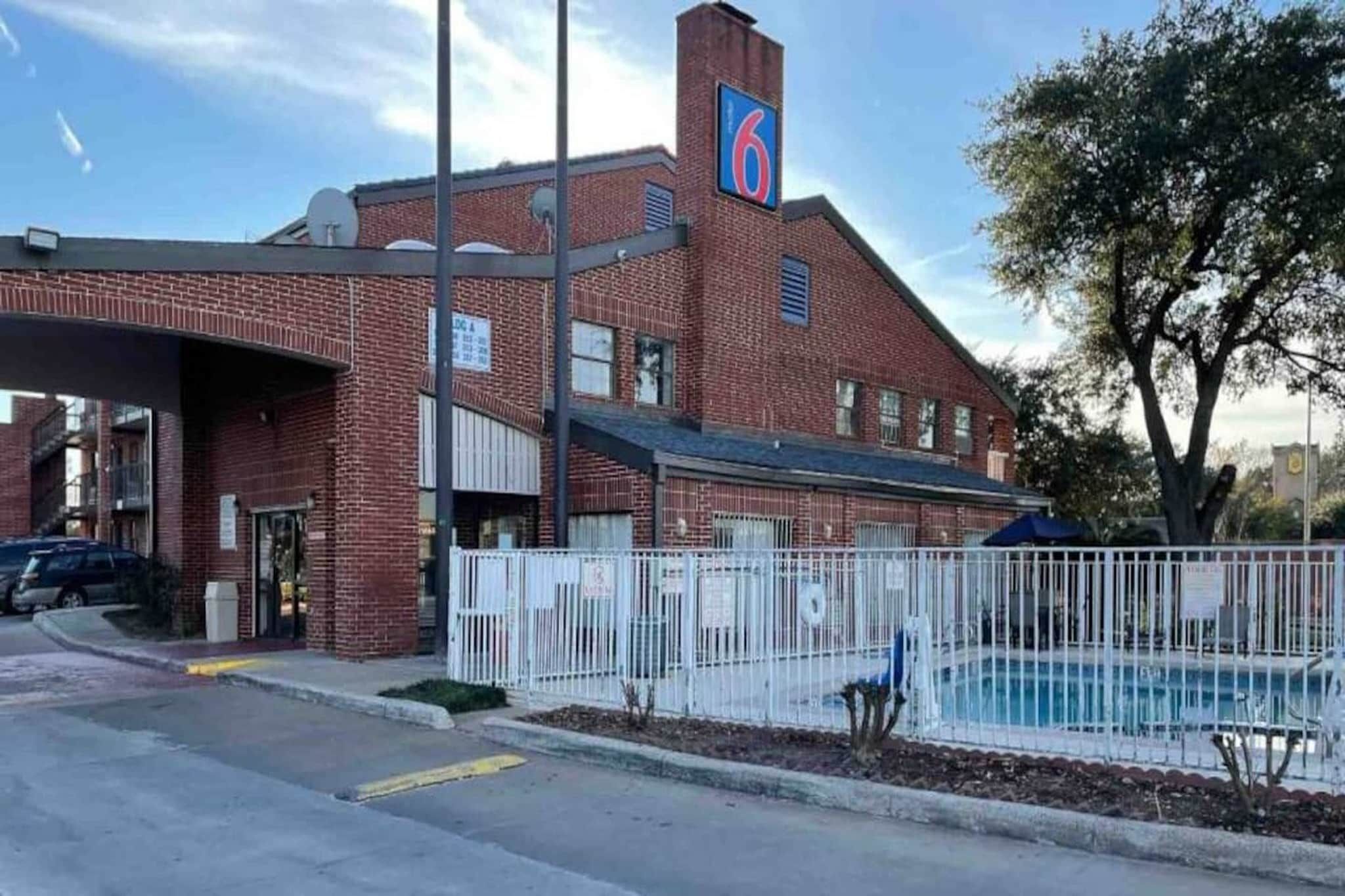 A brick building with a large sign on the roof, a fenced pool area, and a parking lot.
