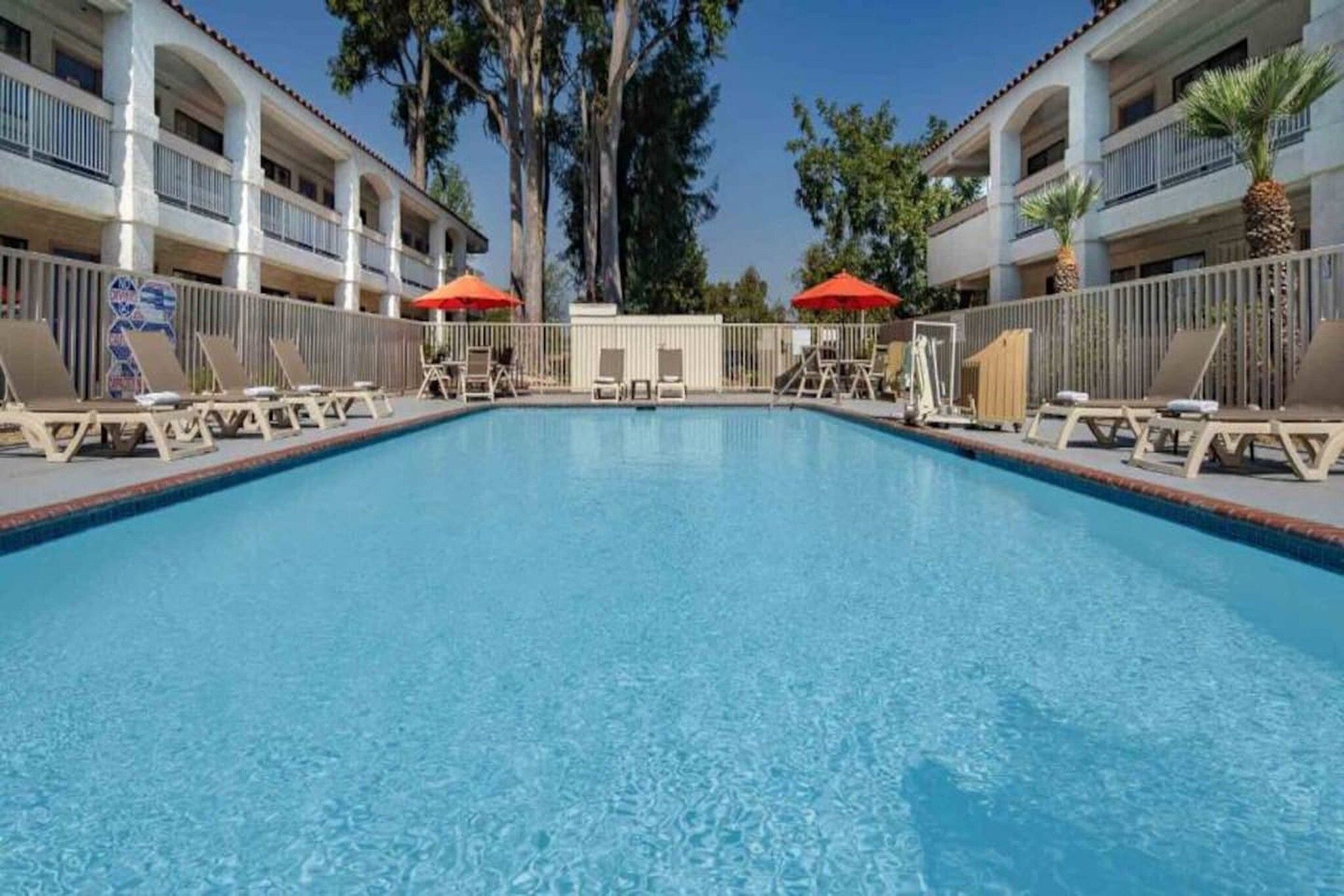 A rectangular swimming pool with lounge chairs and umbrellas, surrounded by a fence, and adjacent to a multi-story building with balconies.