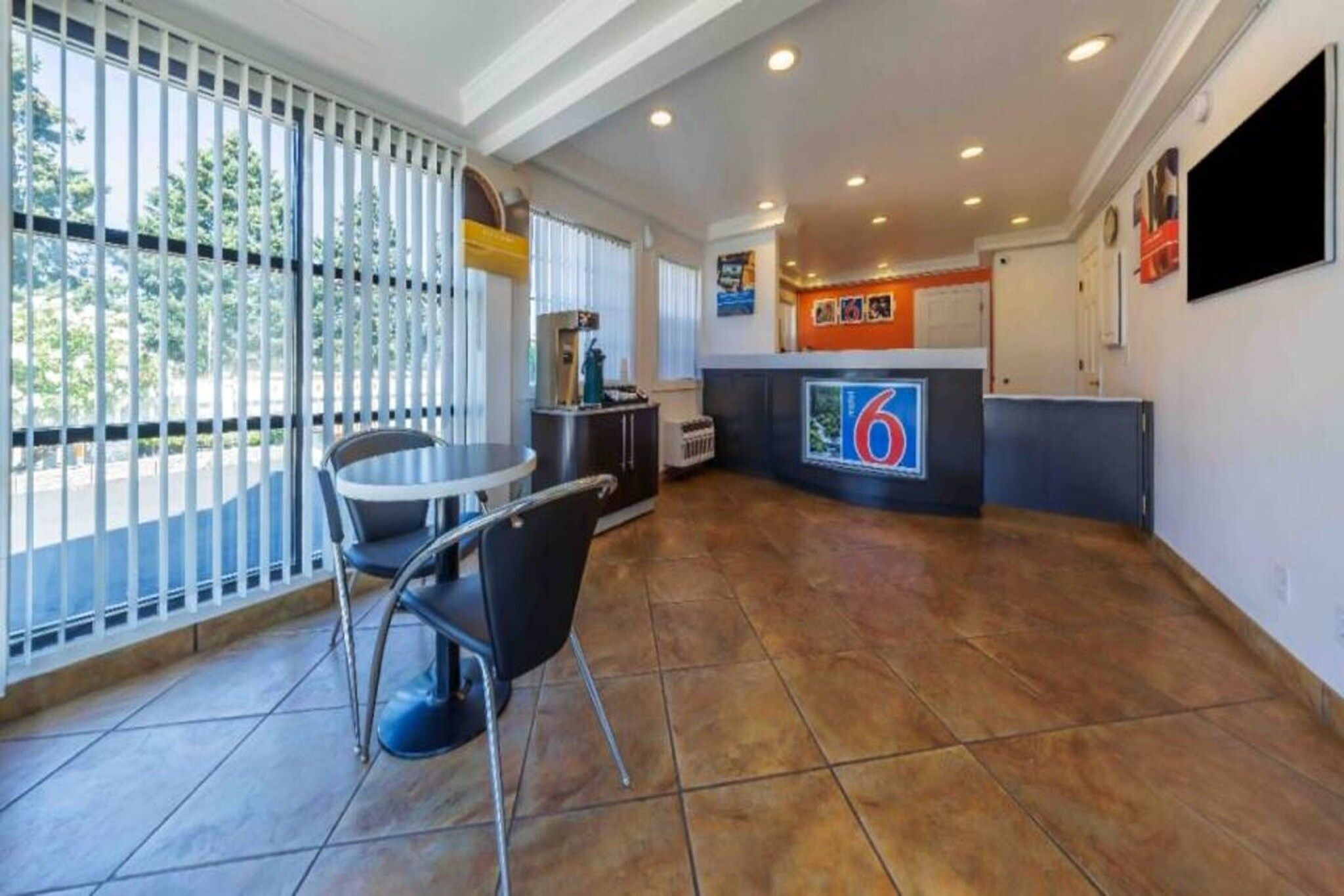 Vestíbulo de hotel con mostrador de recepción, zona de comedor con mesa y sillas, y amplias ventanas con persianas.