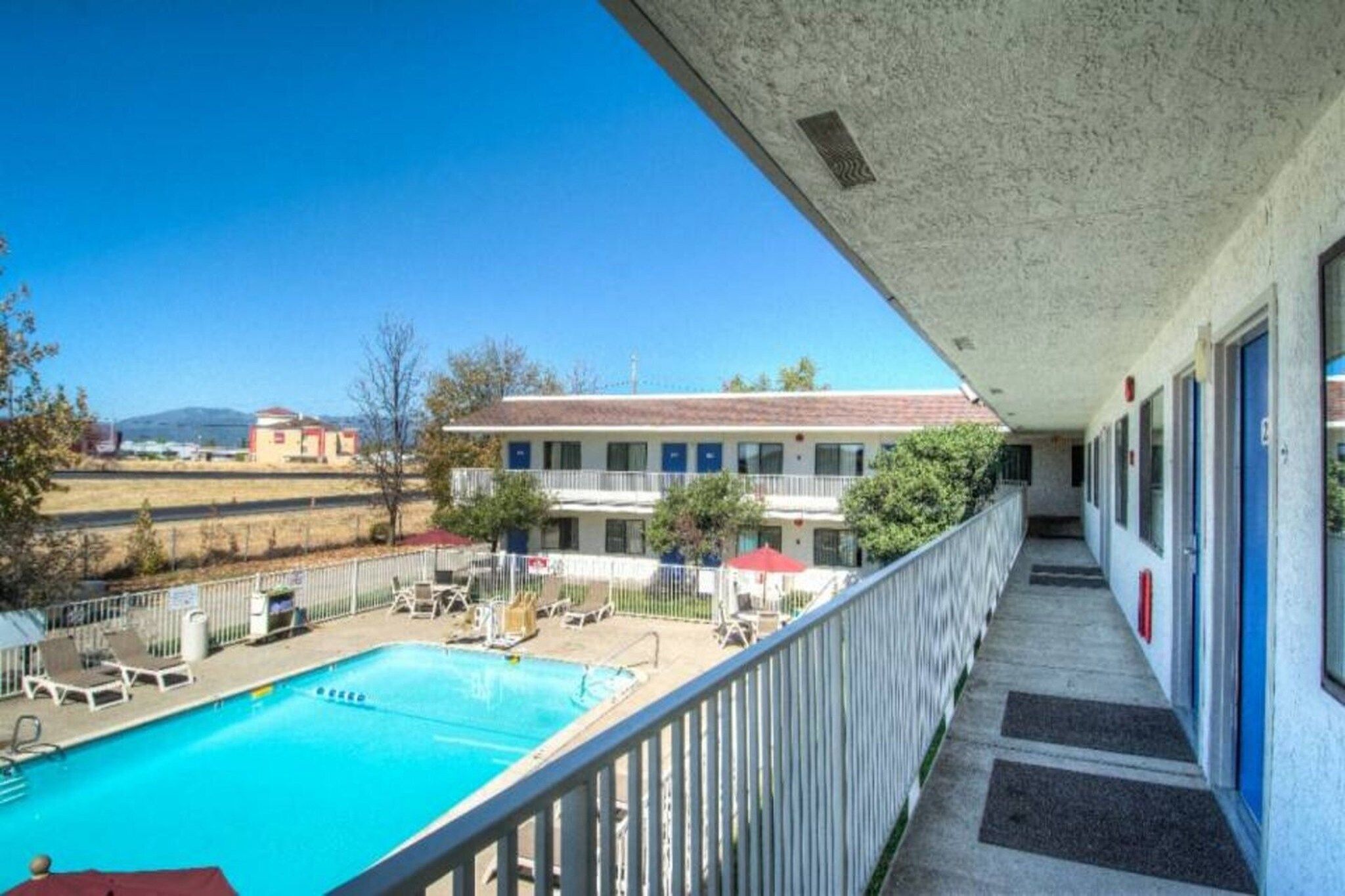 Un balcón de hotel con piscina, mobiliario exterior y vistas del entorno.