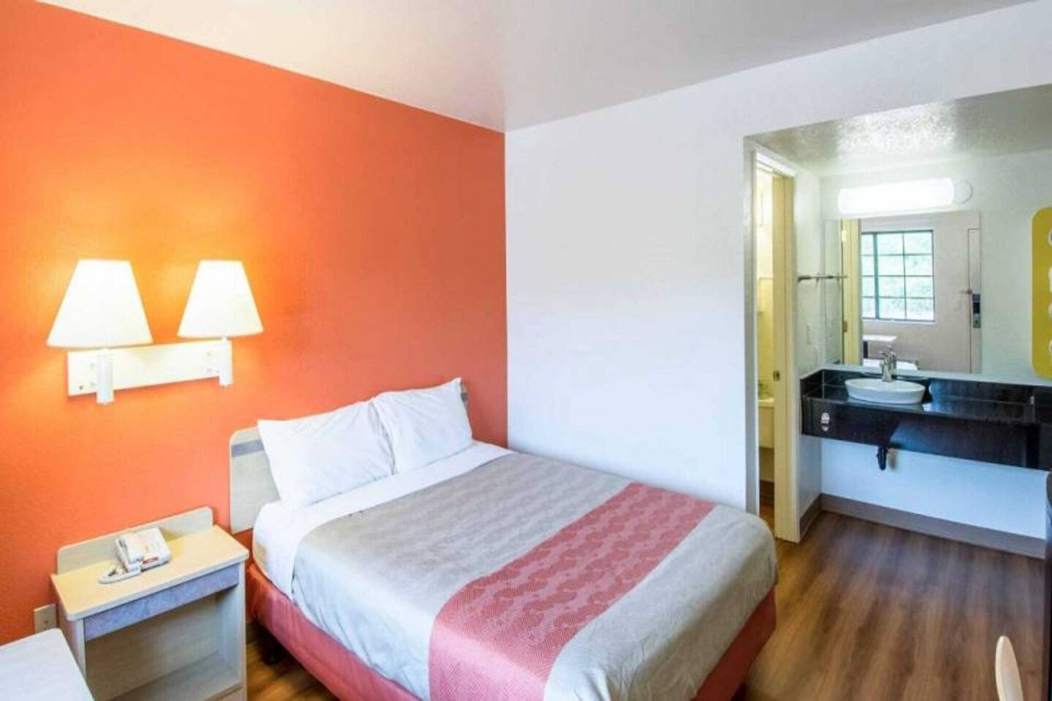 A hotel room with a bed, bedside table, lamp, and a bathroom with a sink and mirror.