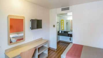 A compact hotel room with a bed, a desk, a chair, a television, and a bathroom visible through an open door.