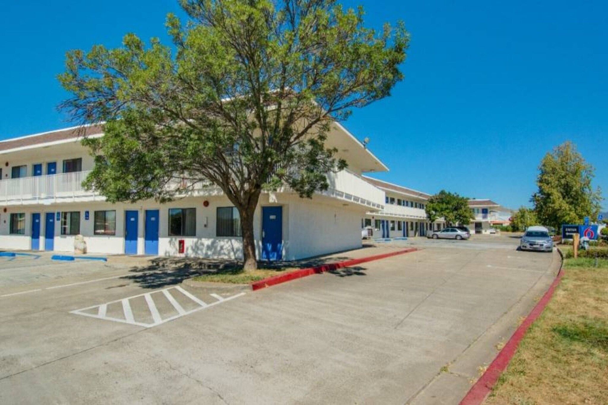 A motel with a parking lot, a tree in front, and a clear blue sky.