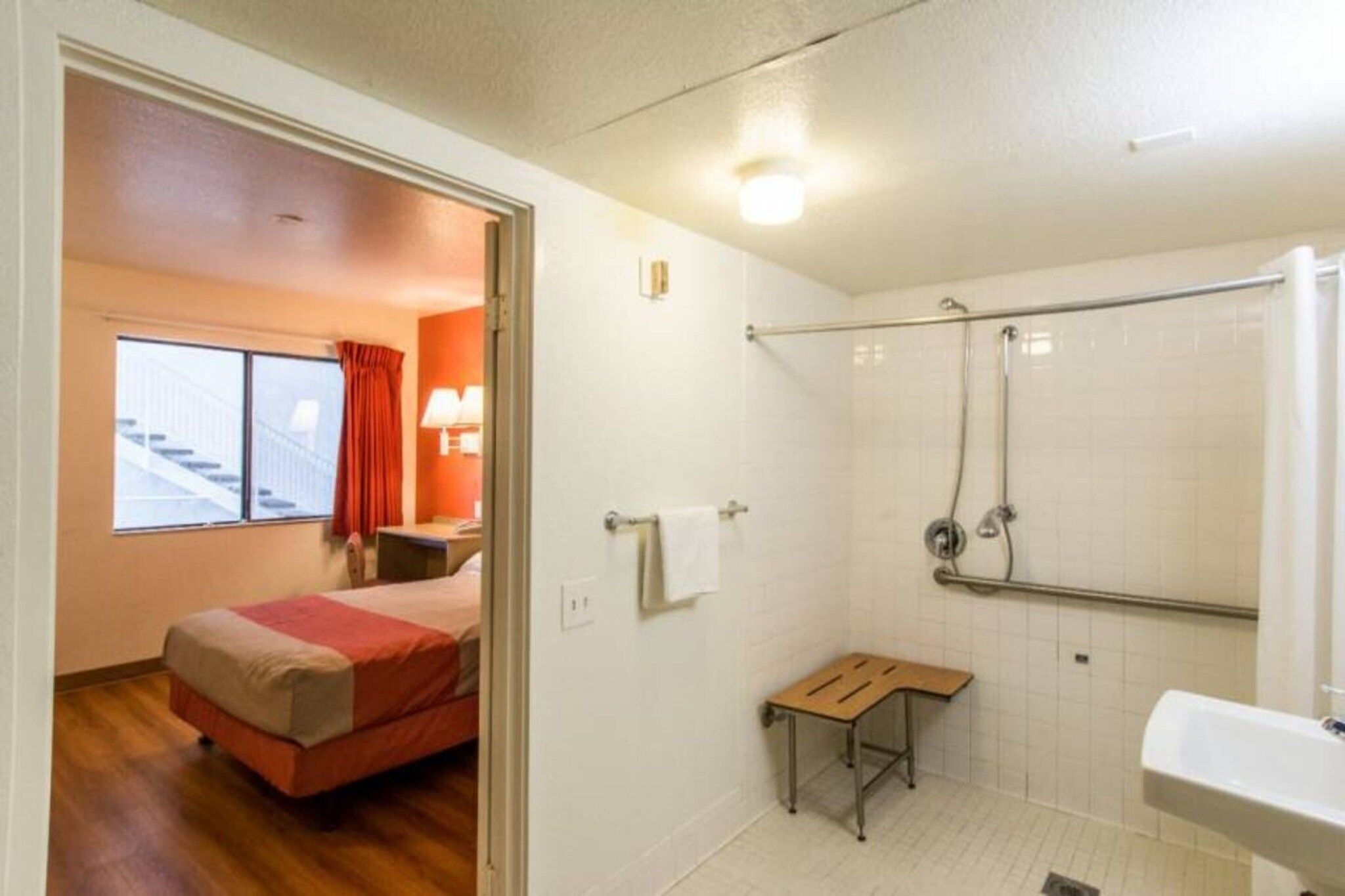 A hotel room with a bed, a small table, a shower area, and a bathroom with a sink.