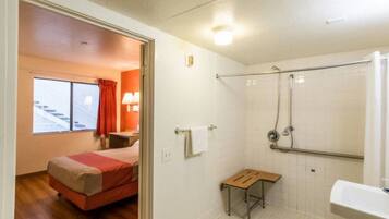 A hotel room with a bed, a small table, a shower area, and a bathroom with a sink.