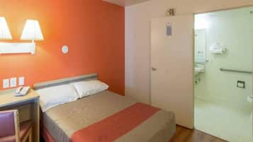 Habitación de hotel con una cama, un escritorio con teléfono, una lámpara de pared y una puerta que da a un baño.