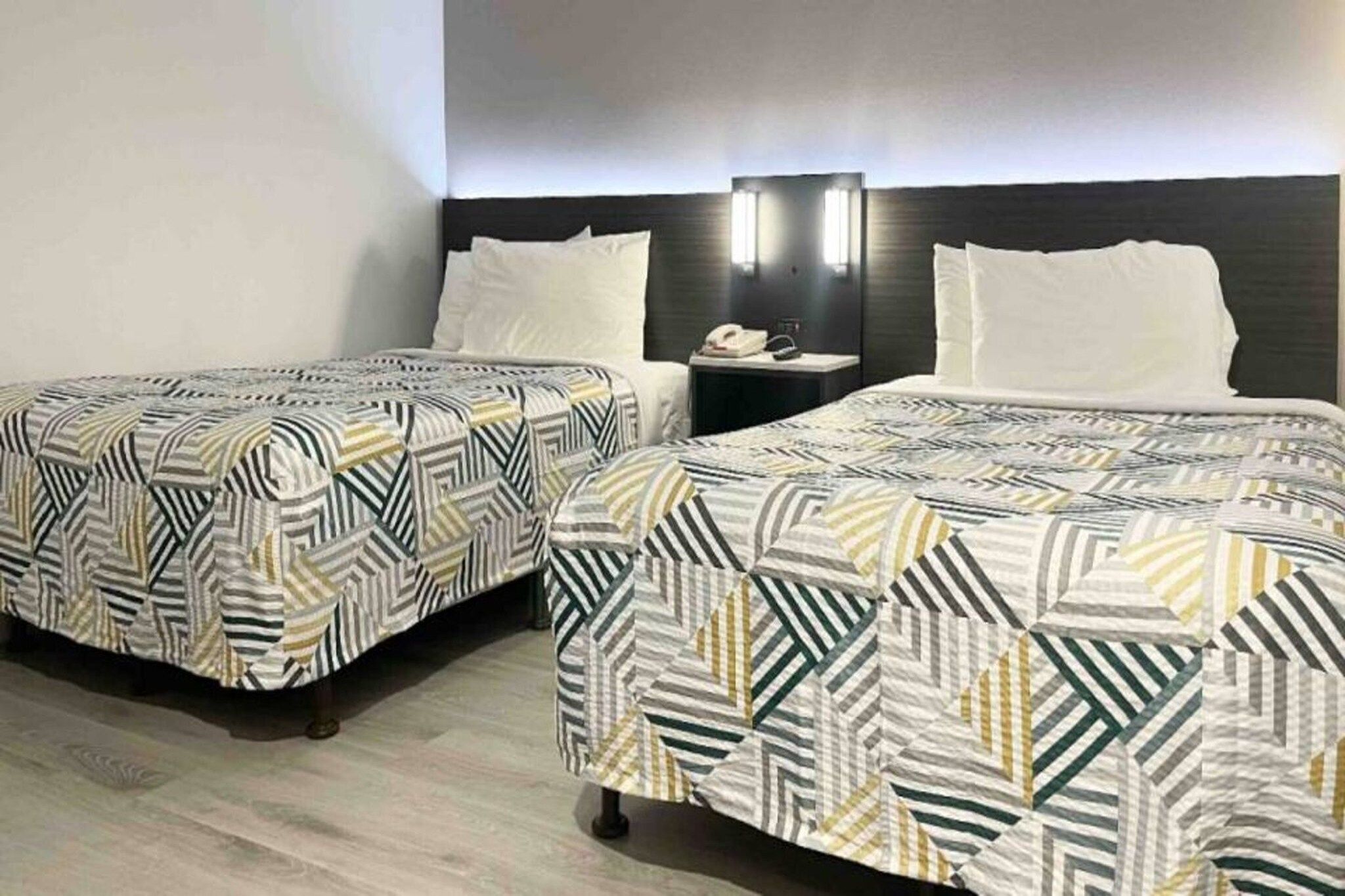 A hotel room with two beds, each with a patterned bedspread, a bedside table with a lamp, and a wall-mounted TV.