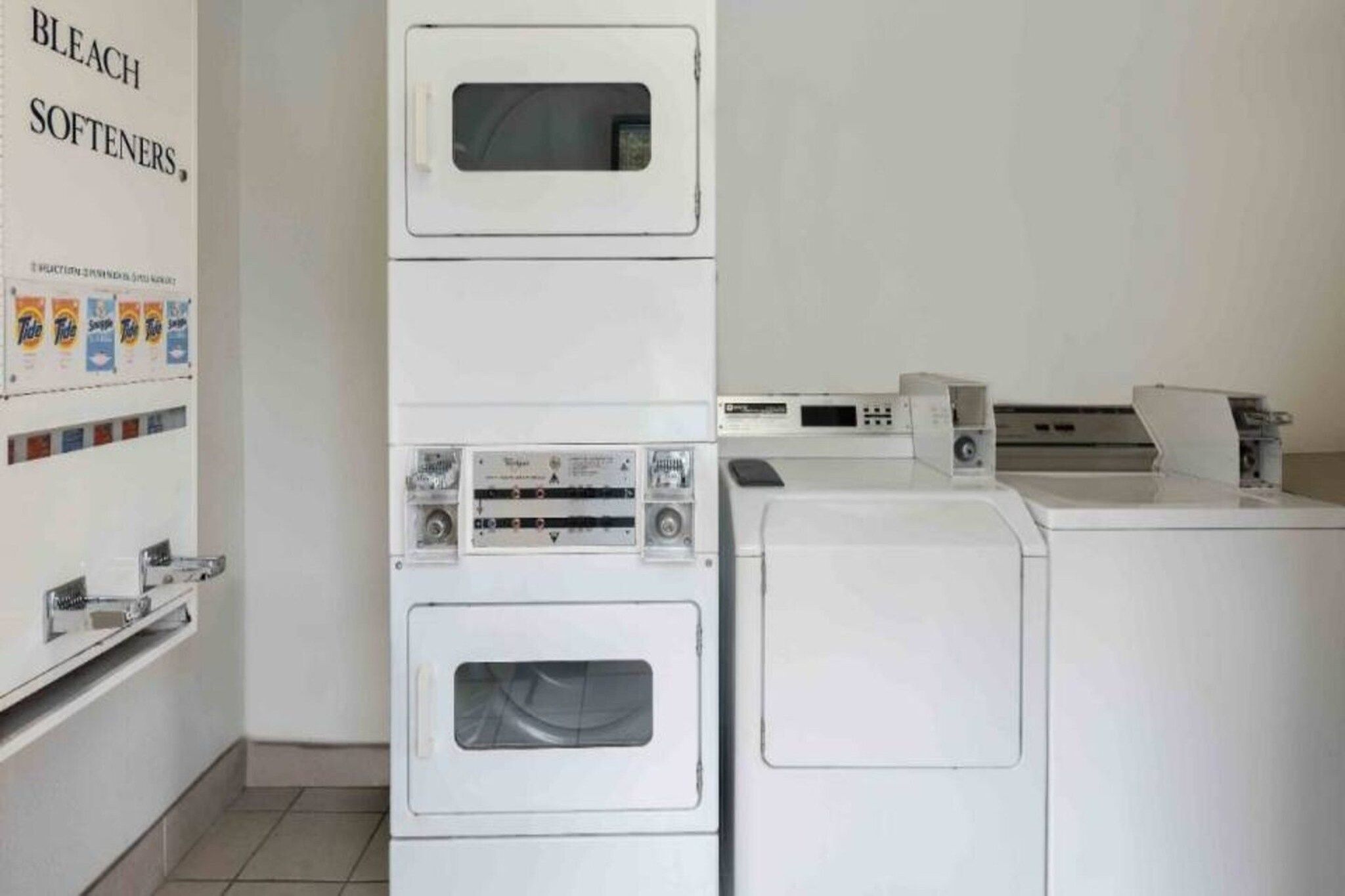 A laundry area with stacked washing machines and dryers, a wall-mounted washing machine, and a shelf with detergent bottles.