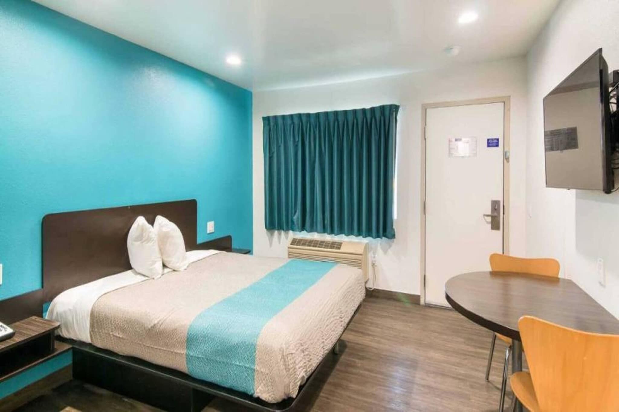 Quarto de hotel com cama, escrivaninha, cadeira, televisão e janela com cortinas.