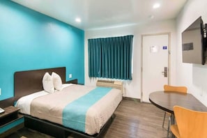 Quarto de hotel com cama, escrivaninha, cadeira, televisão e janela com cortinas.
