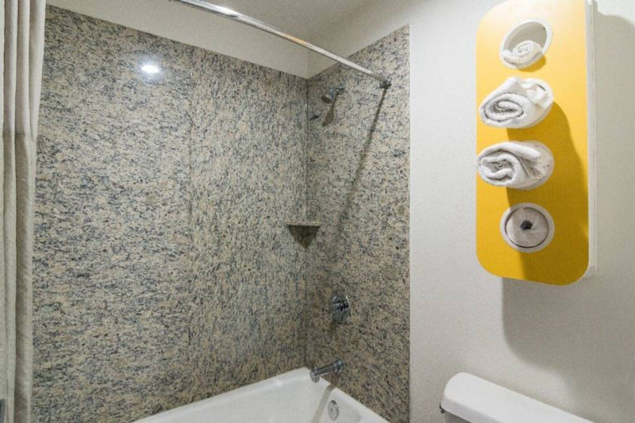 A bathroom with a granite shower wall, a bathtub, and a towel dispenser with three towels.