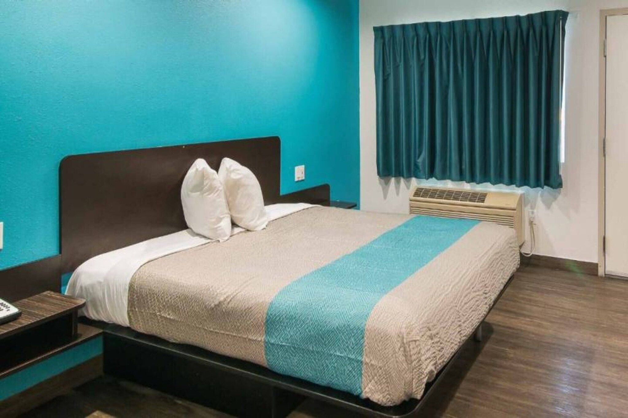 A hotel room with a bed, a nightstand, a window with curtains, and a wall heater.