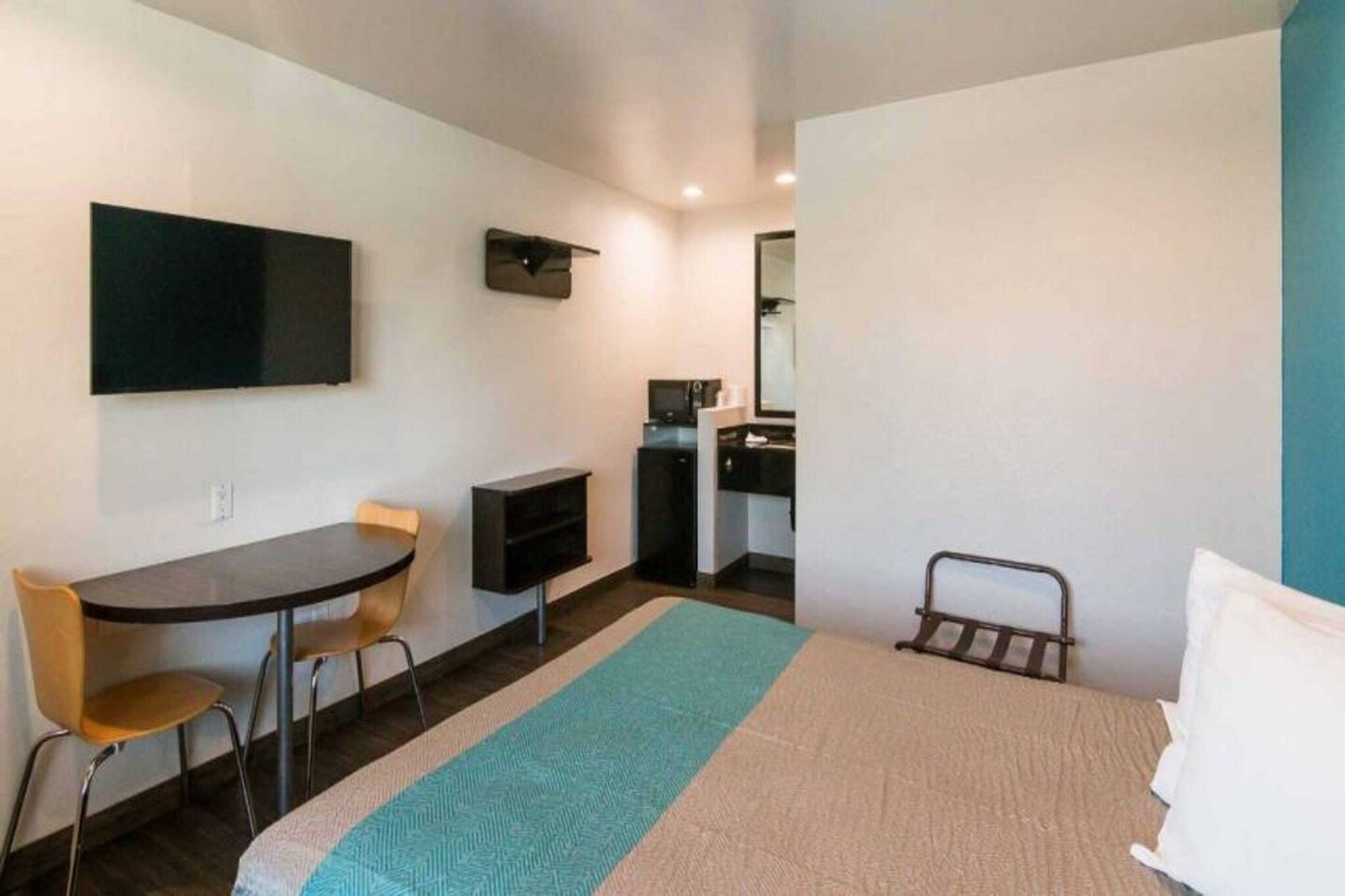 A compact hotel room with a bed, a desk, a chair, a TV, and a small kitchenette.