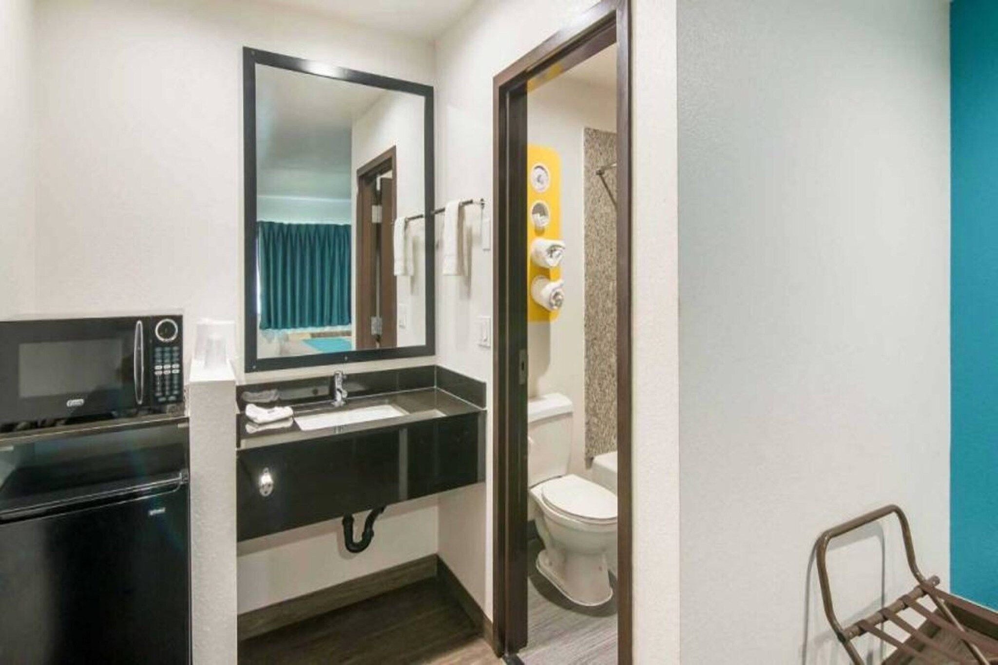 A compact hotel bathroom with a sink, mirror, toilet, and a microwave.