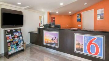 A hotel reception area with a counter, a television mounted on the wall, a magazine rack, and a sign with a sunset image and text.