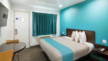 A hotel room with a bed, a desk, a chair, a window with curtains, and a wall with a light switch.