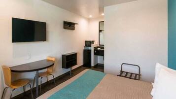 Quarto de hotel pequeno com cama, escrivaninha, cadeira, televisão e uma kitchenette.