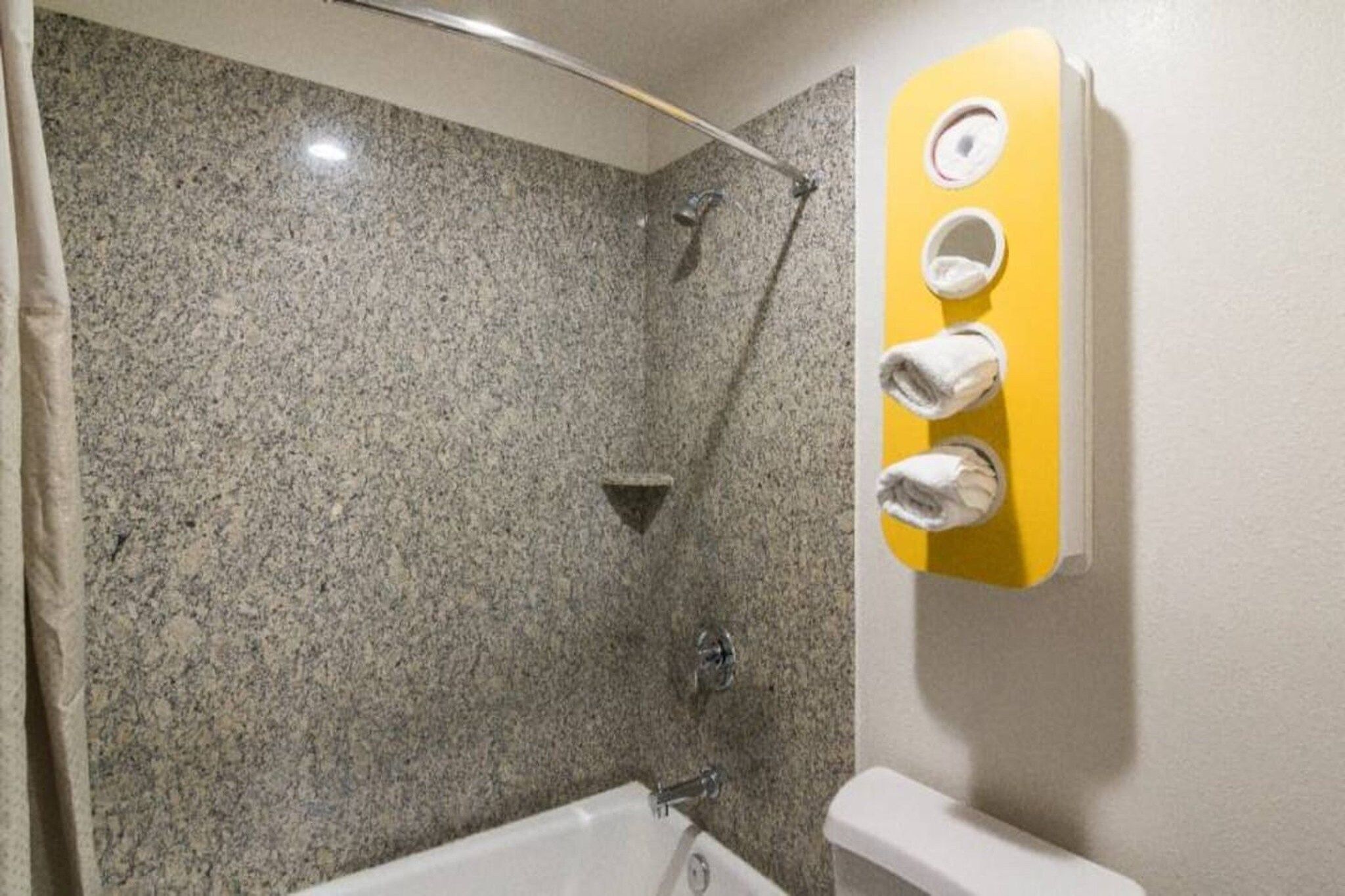 A bathroom with a granite shower wall, a white bathtub, and a yellow towel rack with three towels.