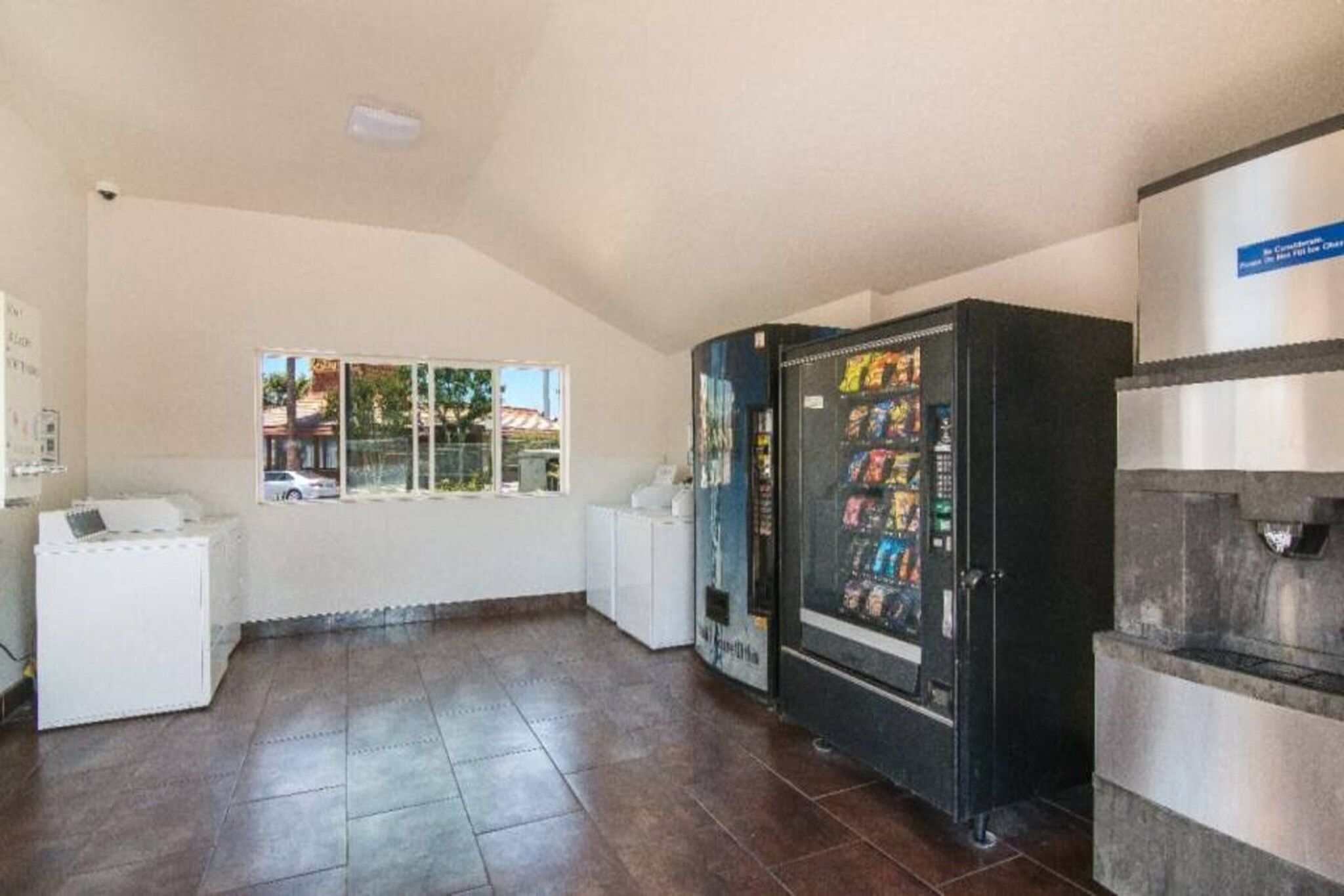A room with a vending machine, a white refrigerator, and a window with a view of buildings.