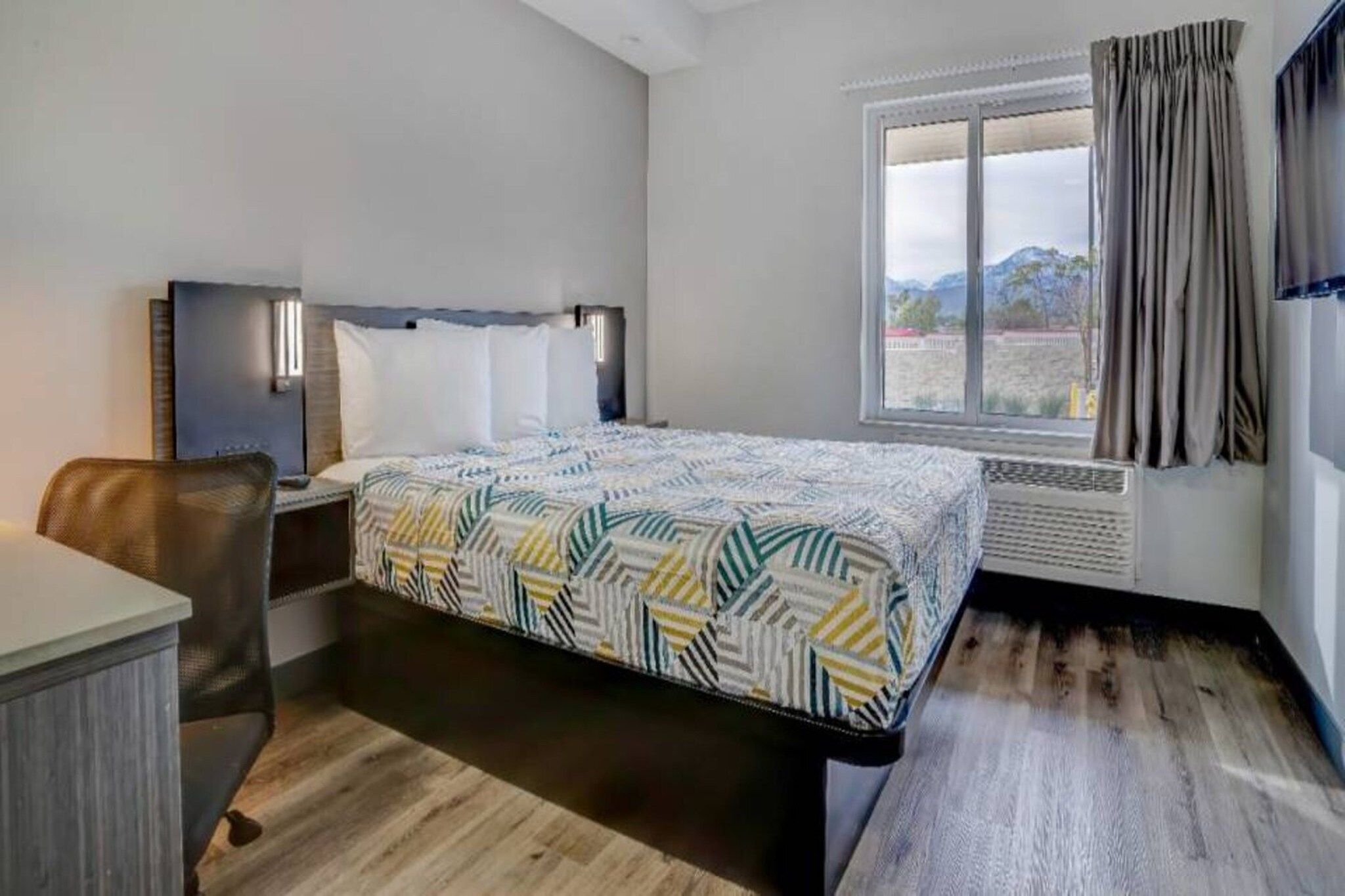 Ein Hotelzimmer mit einem Bett, einem Schreibtisch, einem Stuhl, einem Fenster mit Vorhängen und Blick ins Freie.