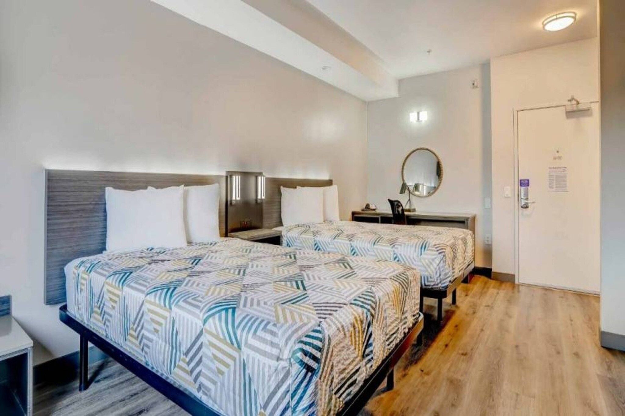 A hotel room with two beds, a bedside table, a mirror, and a door.