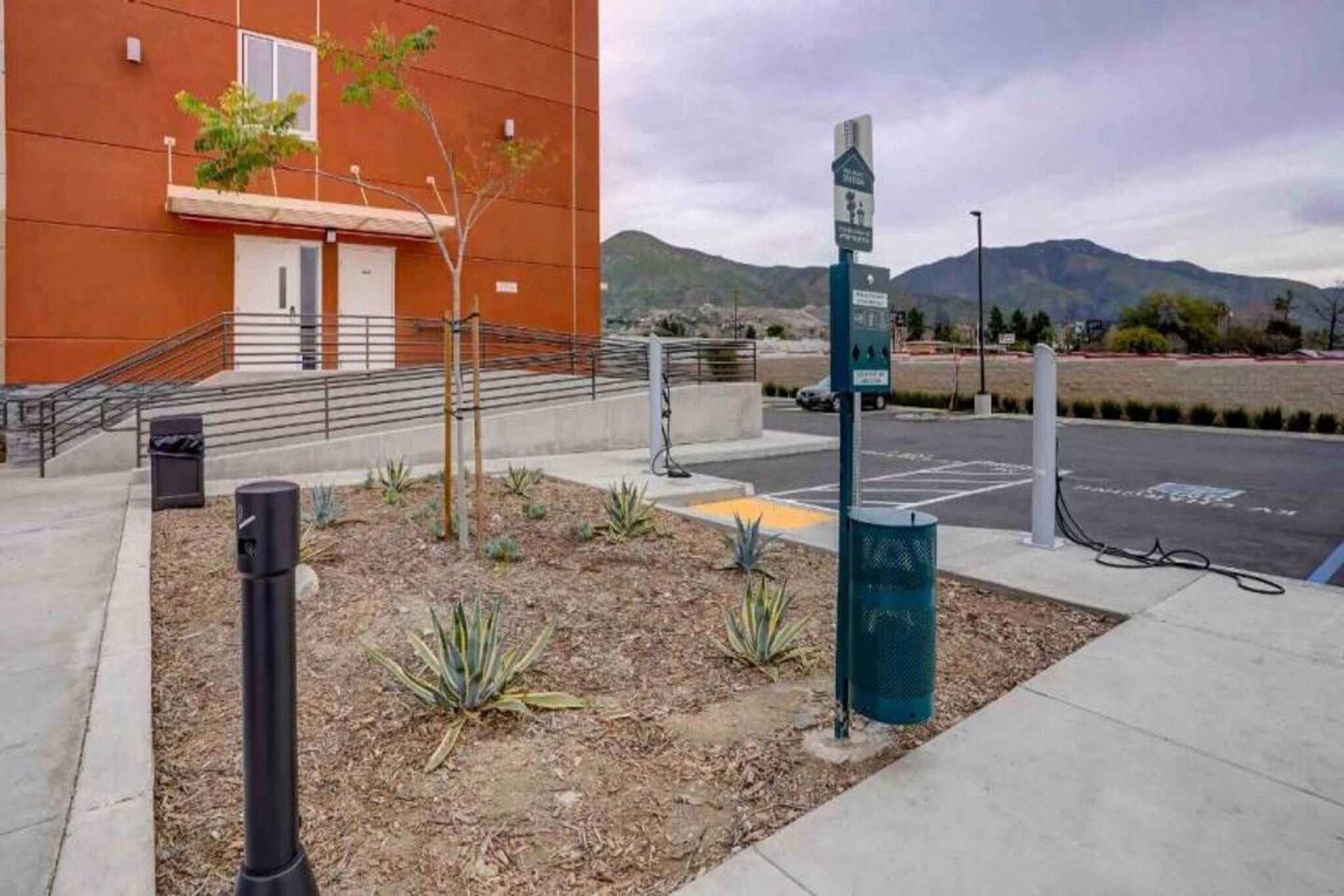 Un área de estacionamiento con estaciones de carga para vehículos eléctricos, un edificio de fachada roja y un paisaje montañoso al fondo.