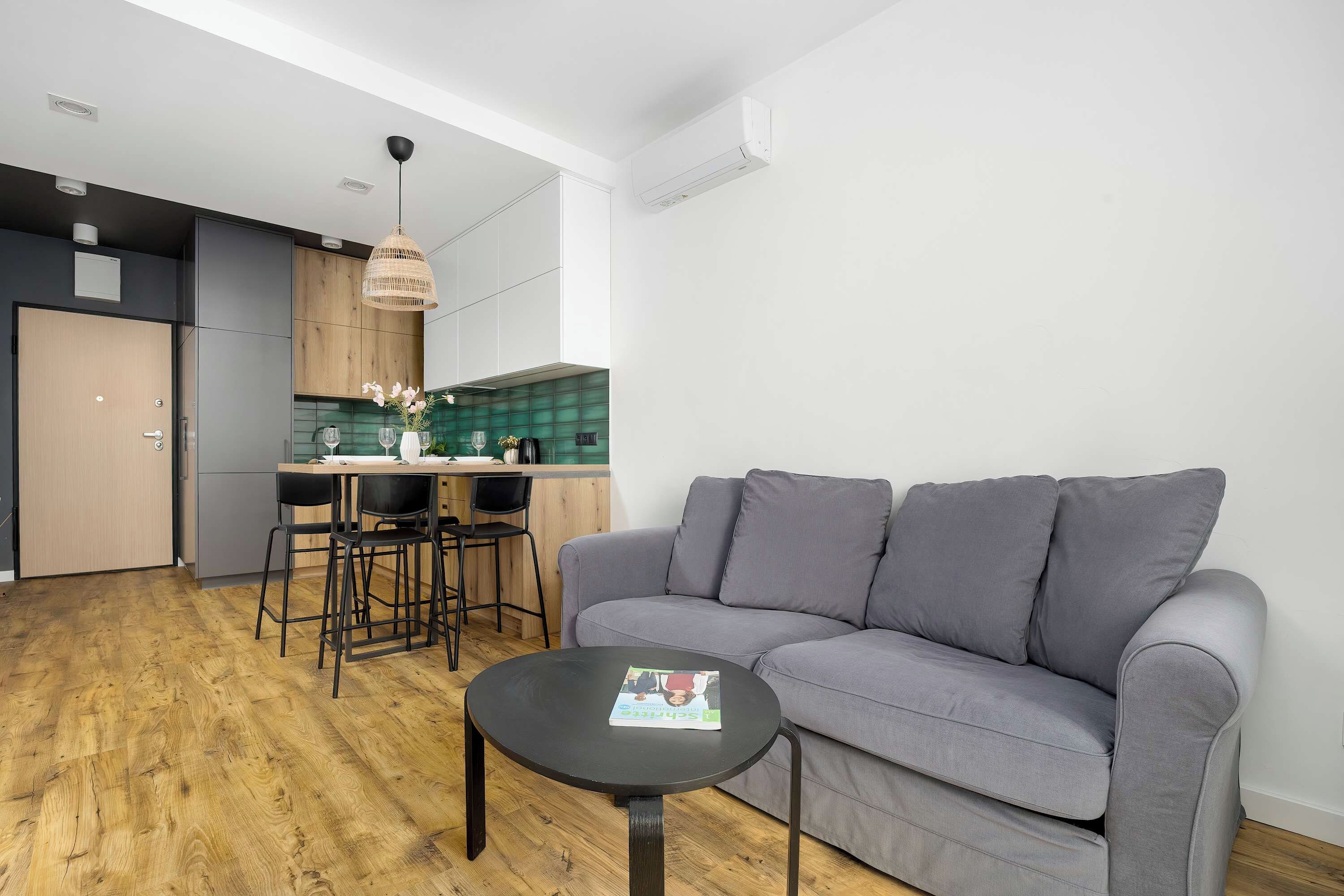 Ein modernes Wohnzimmer mit einem grauen Sofa, einem kleinen runden Tisch und einer Küchenzeile mit Esstisch und Stühlen.