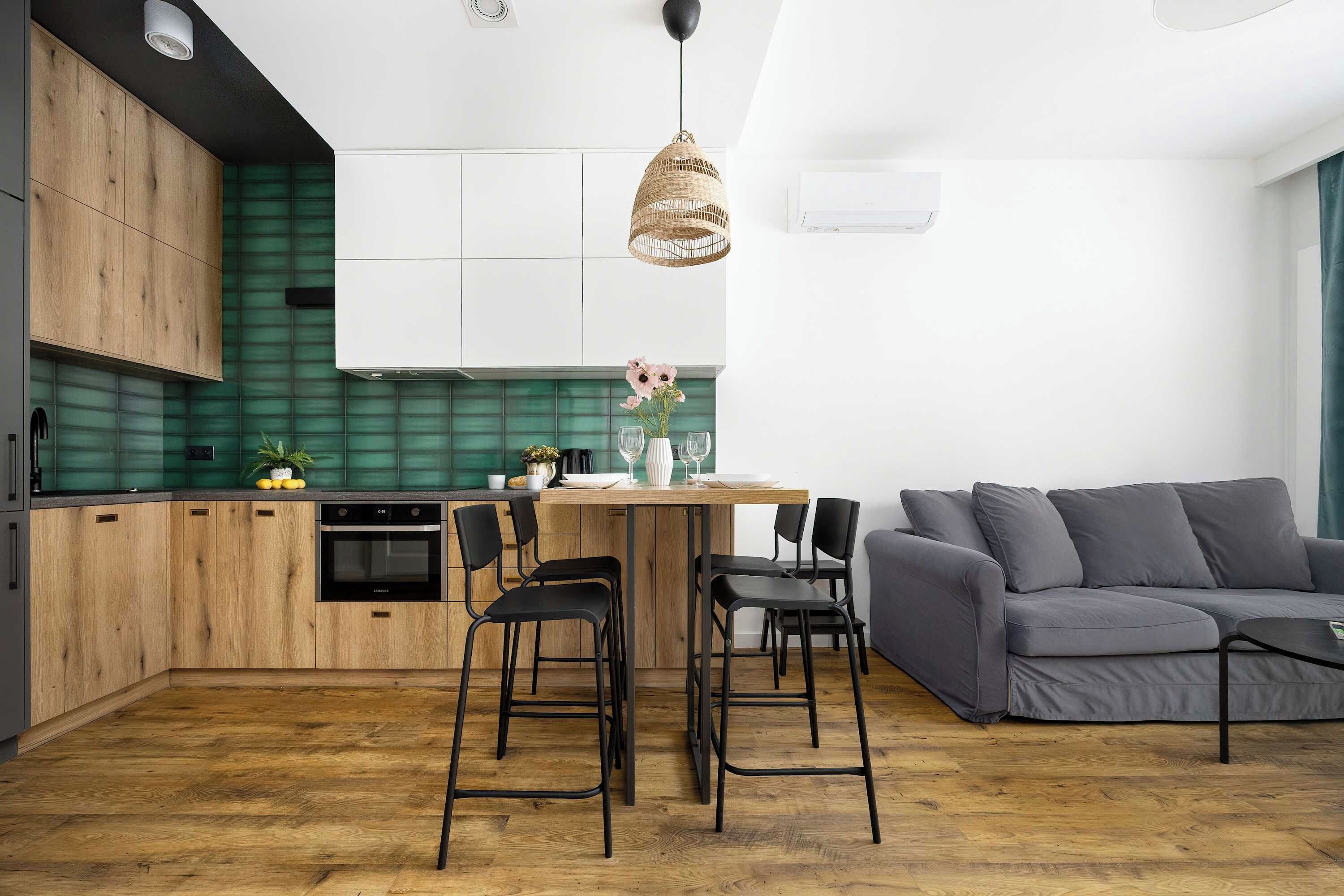 A modern kitchen with wooden cabinets, a green backsplash, and a dining area with a grey sofa and black chairs.