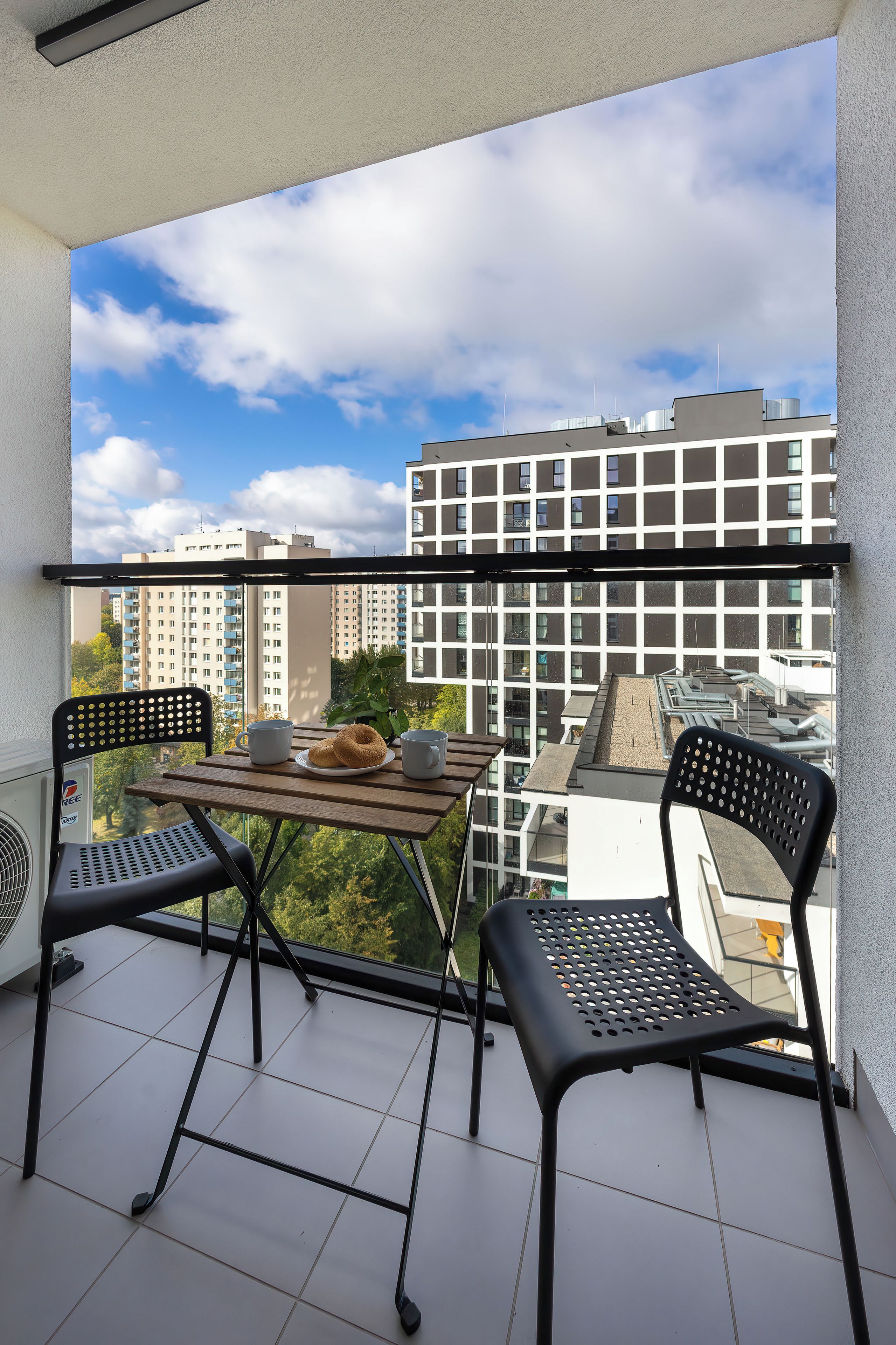 Un balcón con una mesa, dos sillas, una planta en maceta y vistas a un conjunto de edificios.