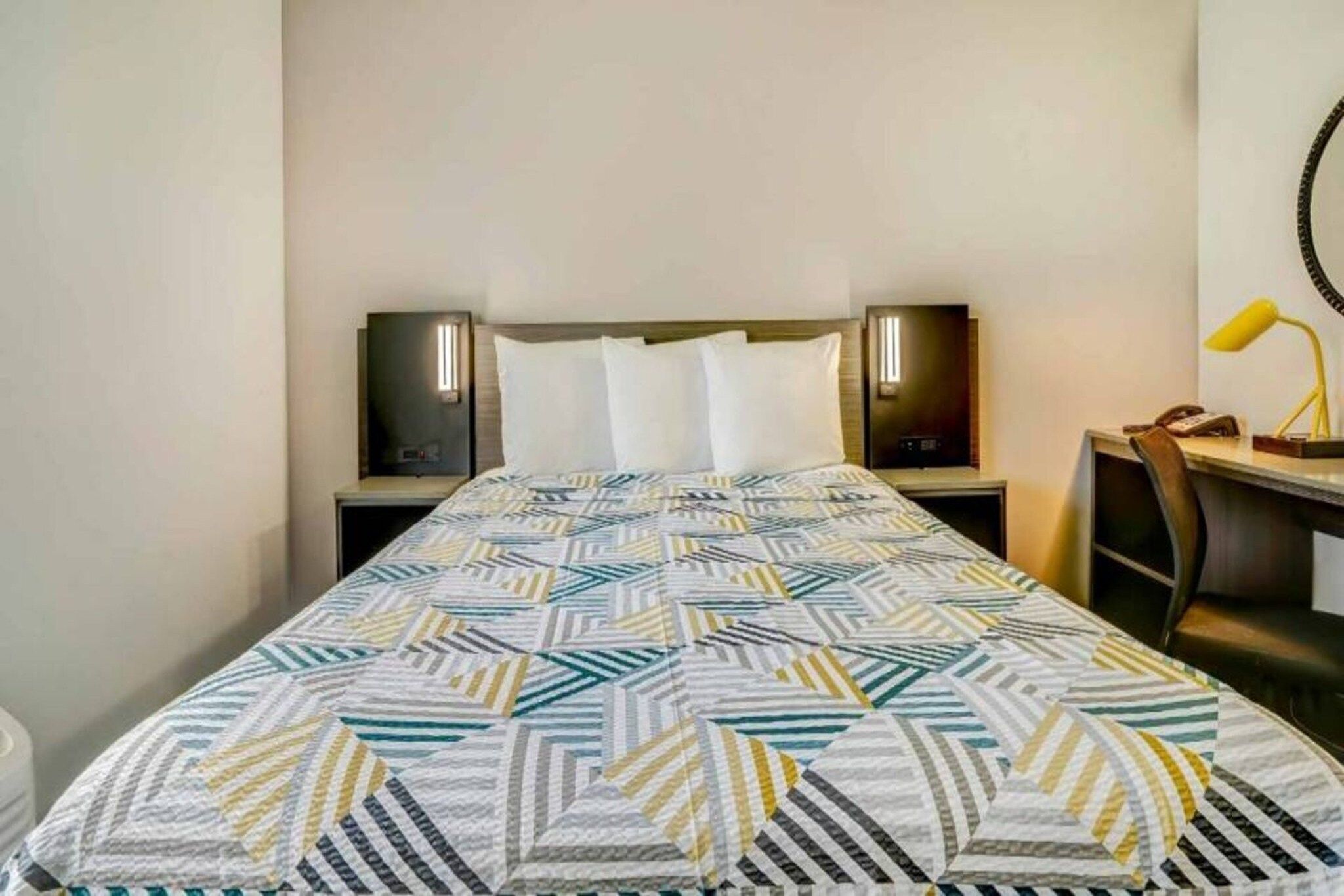 A hotel room with a bed, two bedside tables with lamps, a desk with a chair, and a wall-mounted lamp.