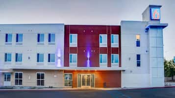 A multi-story hotel with a prominent '6' sign, a central entrance with glass doors, and a parking lot in front.