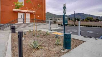 Un área de estacionamiento con estaciones de carga para vehículos eléctricos, un edificio de fachada roja y un paisaje montañoso al fondo.