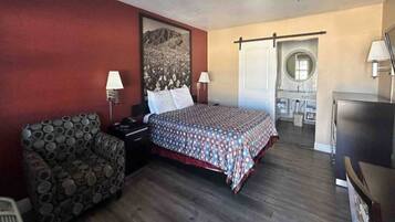 A hotel room with a bed, a chair, a desk, a TV, and a bathroom with a mirror and sink.