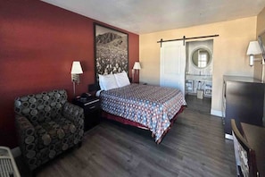 Een hotelkamer met een bed, een stoel, een bureau, een lamp, een spiegel en uitzicht op een balkon.