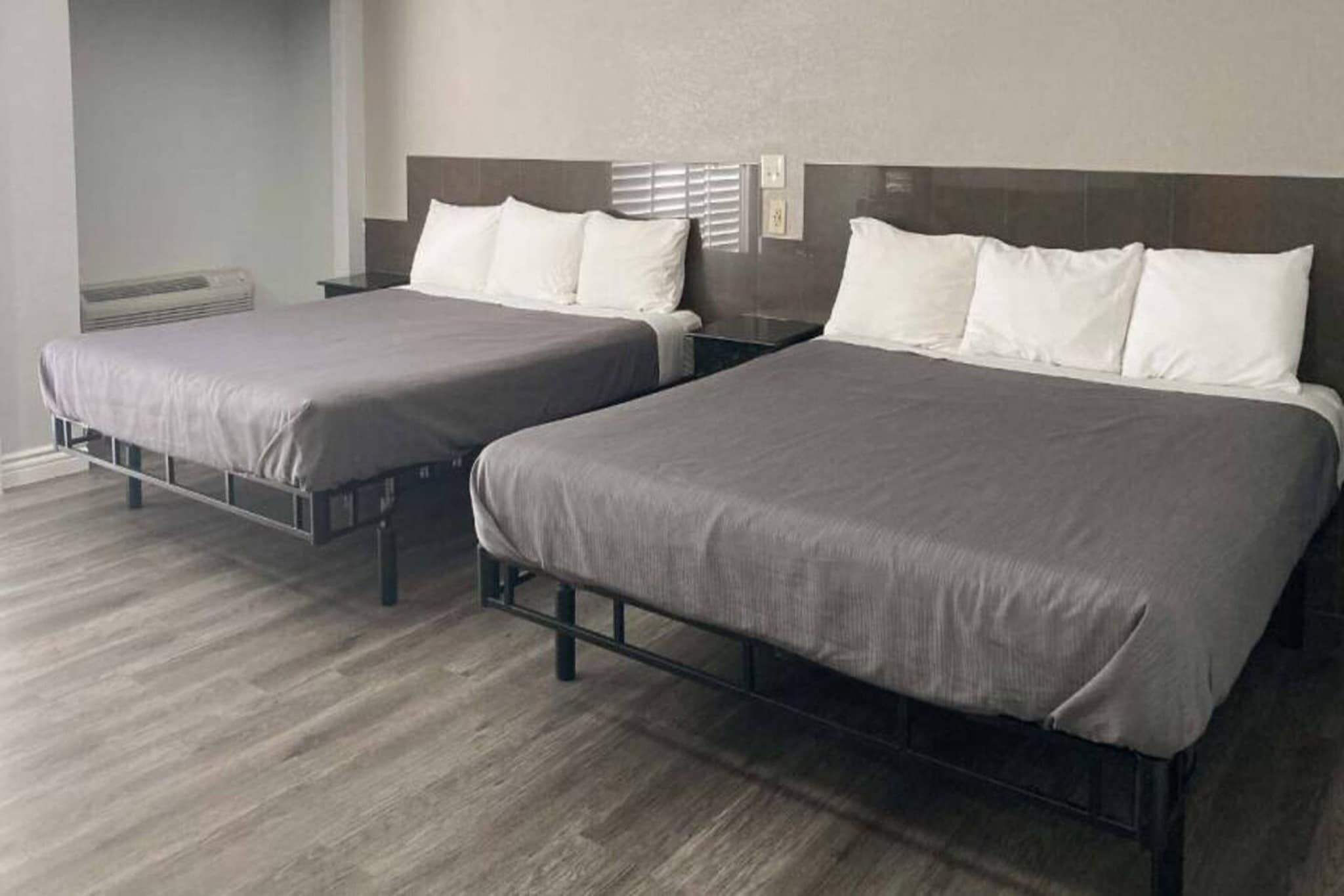 Two twin beds with metal frames and grey bedding, a wooden floor, and a wall with a window and electrical outlets.