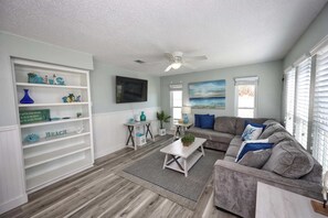 A living room with a grey sofa, a coffee table, a TV, and a bookshelf with decorative items. - Seashell Retreat | Beachfront + Gulf Views (Galveston)