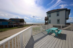 Une terrasse en bois avec une table de pique-nique, une clôture blanche et un bâtiment doté d’un balcon donnant sur la plage.