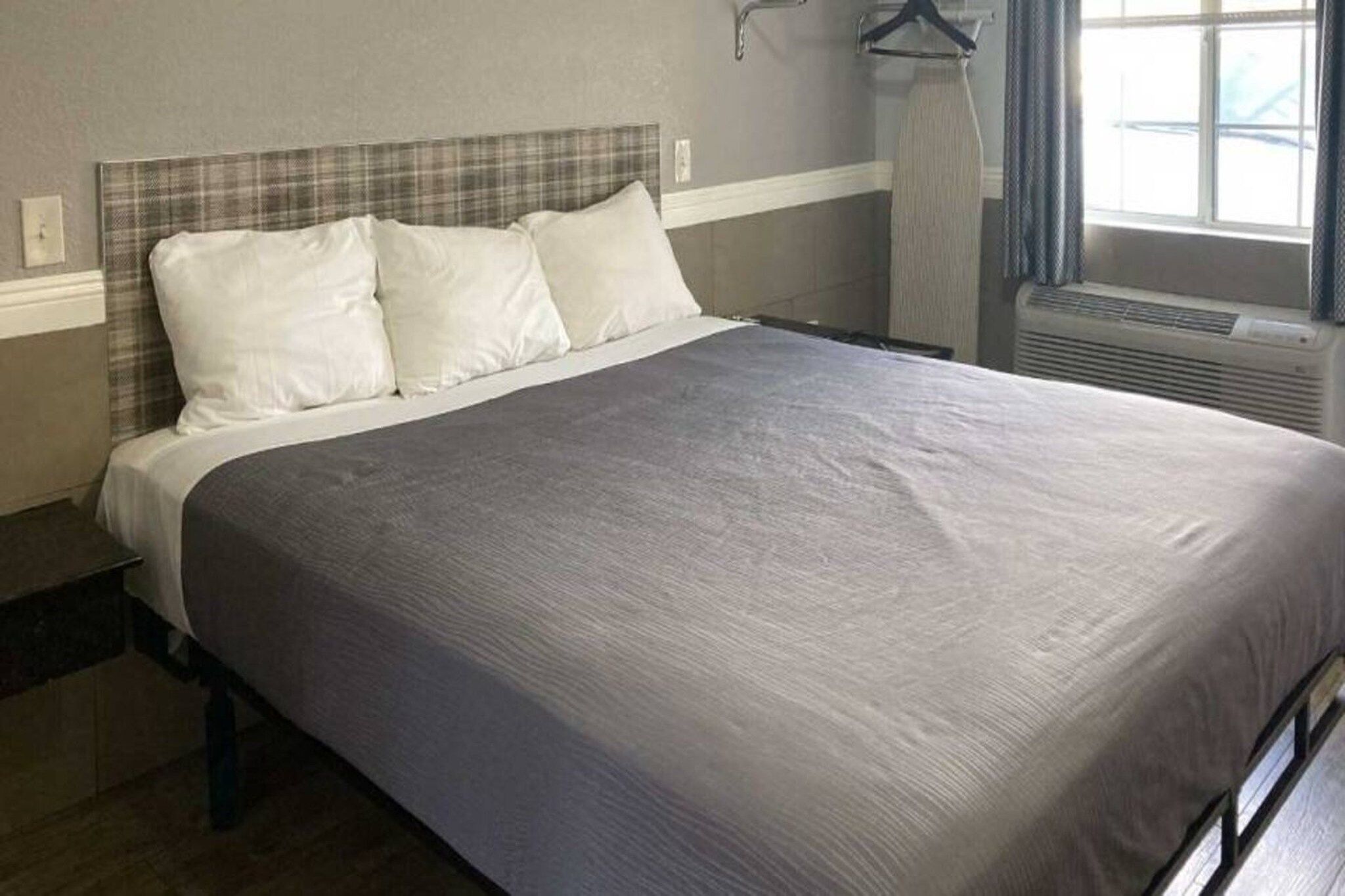 A bed with a grey bedspread and white pillows in a room with a grey headboard and a window with curtains.