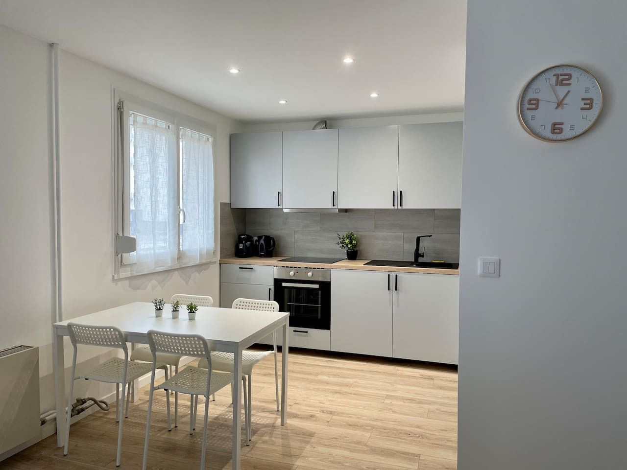 Een moderne keuken met witte kasten, een eettafel met stoelen en een klok aan de muur.