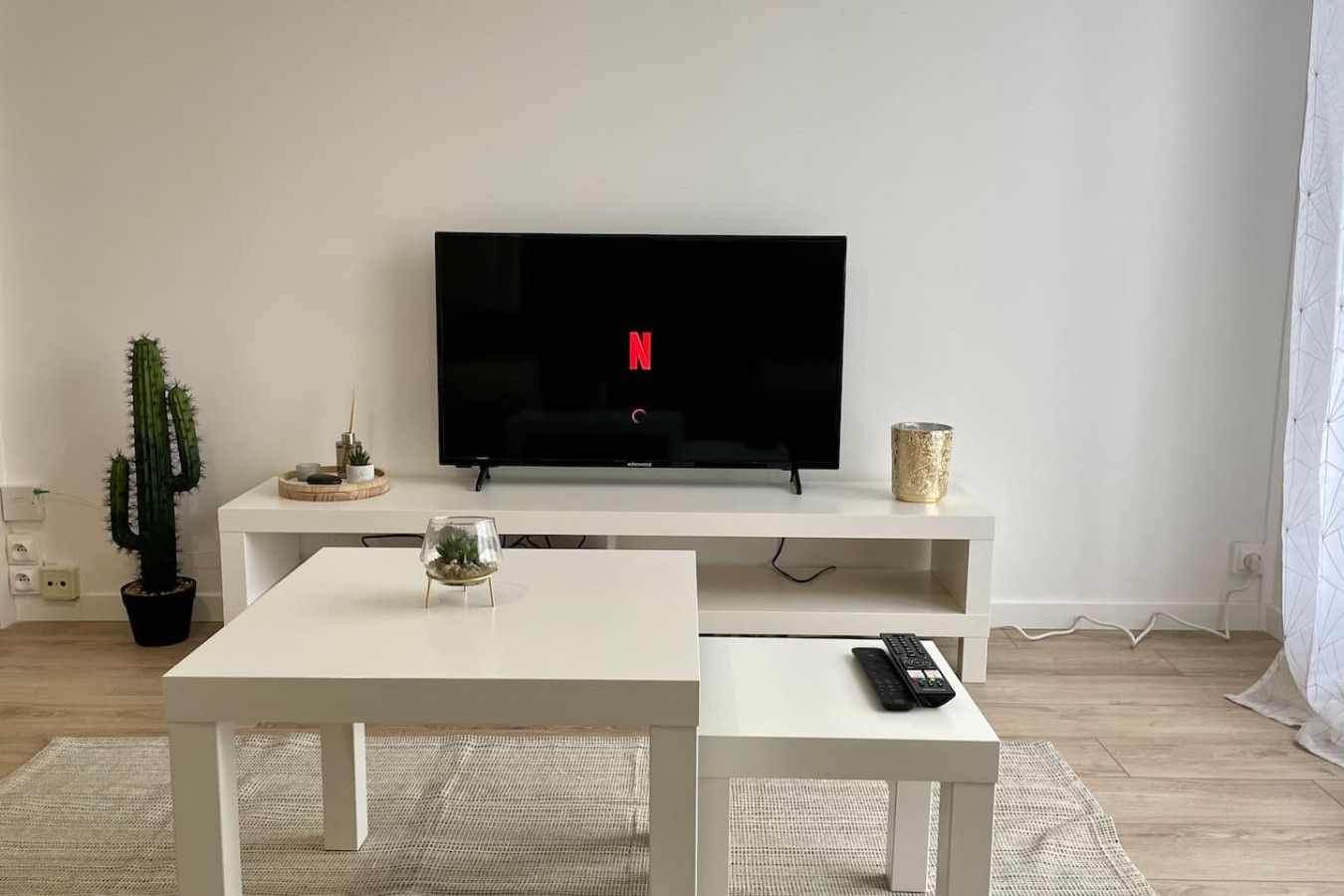 Un salon avec un téléviseur à écran plat posé sur un meuble blanc, une petite table sur laquelle se trouve un récipient en verre, un cactus et une télécommande posée sur le meuble.
