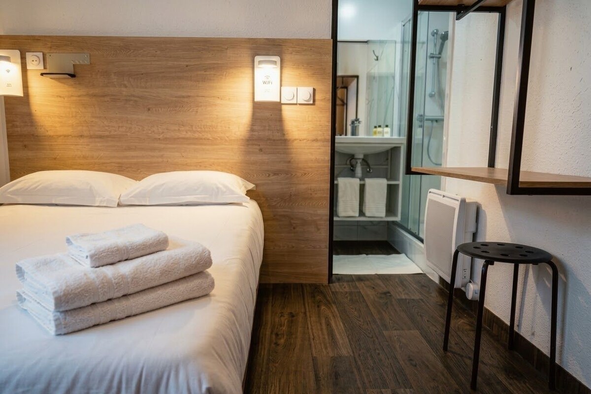 Habitación de hotel con cabecera de madera, cama con ropa de cama blanca, un taburete pequeño y zona de ducha con paredes de vidrio.
