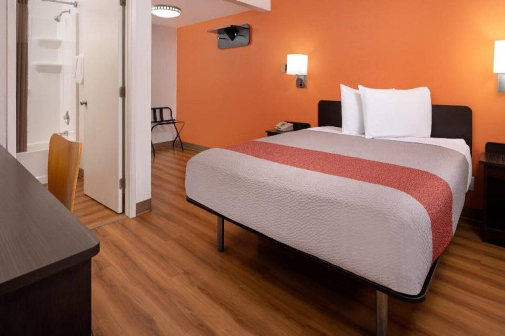 Quarto de hotel com cama, escrivaninha, cadeira, mesa de cabeceira e banheiro com chuveiro e porta-toalhas.
