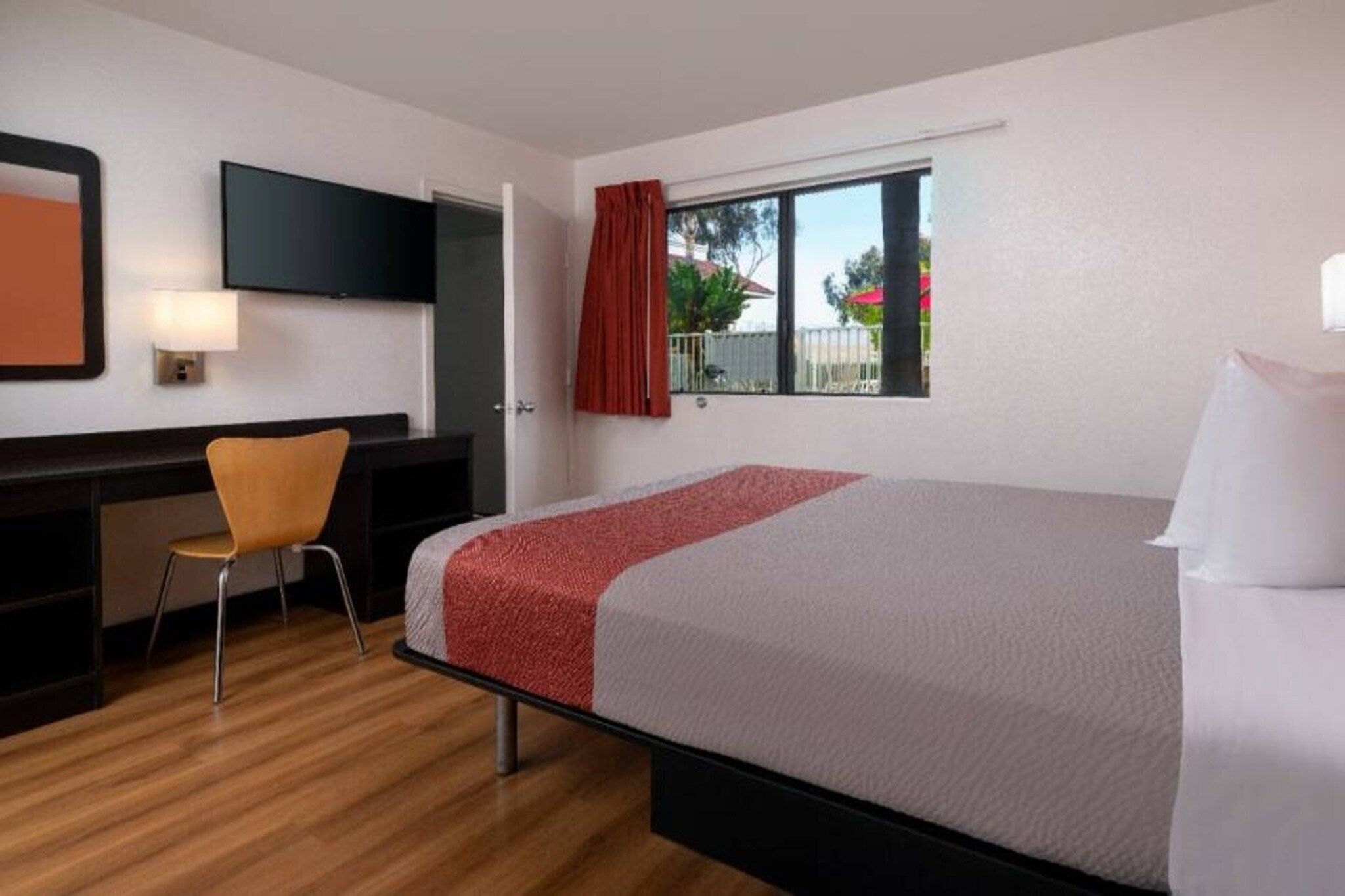Quarto de hotel com cama, escrivaninha, cadeira, televisão e janela com cortinas.