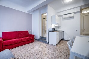 Ein kompakter Raum mit einem roten Sofa, einer kleinen Küchenzeile, einem weißen Esstisch und einer Tür zu einem weiteren Raum.