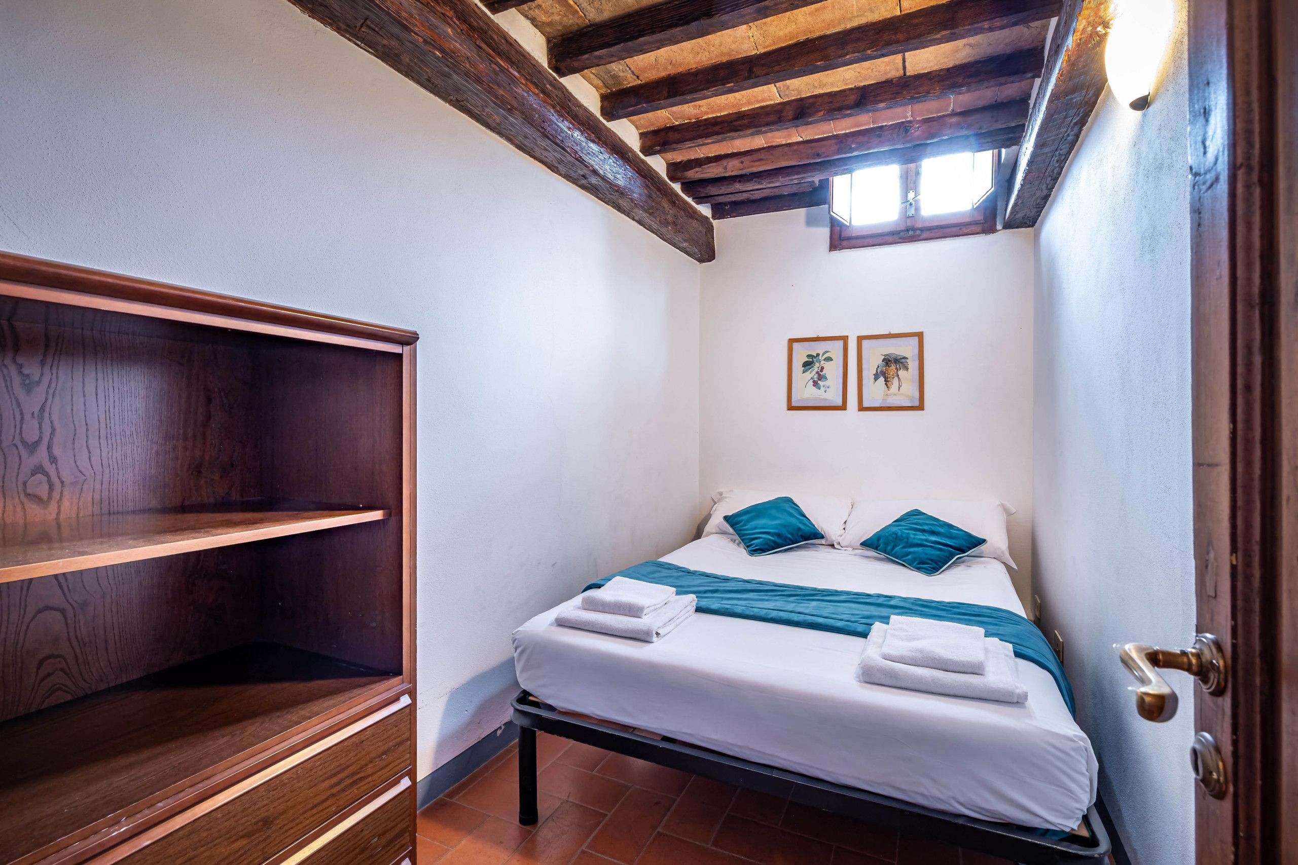 A small, well-lit bedroom with a wooden ceiling, a bed with blue and white bedding, and a wooden wardrobe.